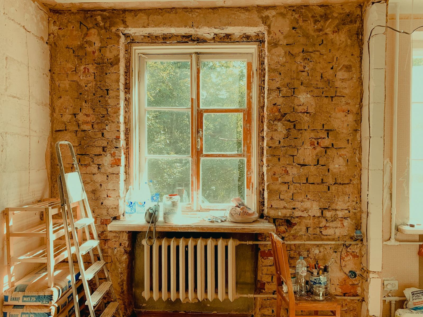 Pièce en cours de rénovation avec fenêtre, radiateur et échelle de peintre